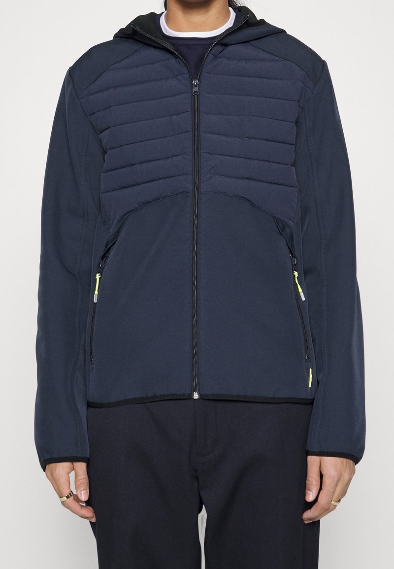 Marinblå jacka med vadderade övre sektioner, dragkedja framtill och sidofickor. Har en huva och ljusa dragkedjeinslag. Slät textur.