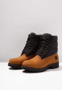 Bottes en daim marron avec une partie supérieure en feutre noir. Caractéristiques : lacets gris, œillets noirs et semelle en caoutchouc noire avec un motif texturé.