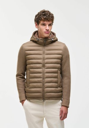 Junger Mann trägt eine braune gesteppte Kapuzenjacke mit gestrickten Ärmeln und weiße Hose, steht vor einem schlichten weißen Hintergrund.