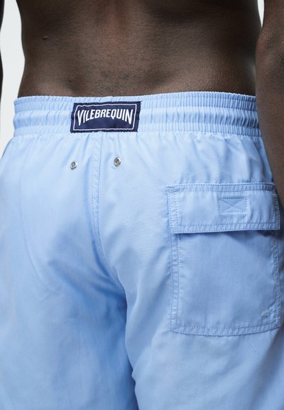Κοντινό πλάνο ενός ατόμου που φοράει ανοιχτό μπλε μαγιό με ελαστική μέση, πίσω τσέπη και ετικέτα της μάρκας Vilebrequin.