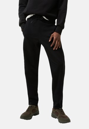 Person trägt schwarze Hose mit Kordelzug, schwarzen Sweater, weißes Unterhemd und braune Wanderschuhe, steht mit Händen nahe den Taschen.