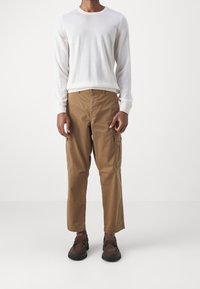 Maglione bianco a maniche lunghe in maglia con scollo rotondo, abbinato a pantaloni cargo beige con tasche laterali e orli risvoltati. Le scarpe marroni completano l'outfit.