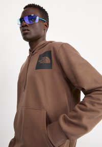 Bruine hoodie met een voorzak en een zwart logo patch. Model draagt blauw-getinte zonnebril met teal accenten. Eenvoudig ontwerp, zachte textuur.