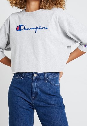 Personne portant un sweat court gris clair avec un logo Champion bleu et un jean bleu taille haute, mains posées sur les hanches.
