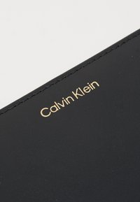 Portefeuille en cuir noir avec un logo doré "Calvin Klein", présentant des coutures visibles le long des bords et une texture lisse.
