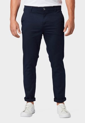 Uomo che indossa pantaloni slim fit blu navy risvoltati e sneakers bianche, in piedi davanti a uno sfondo semplice.