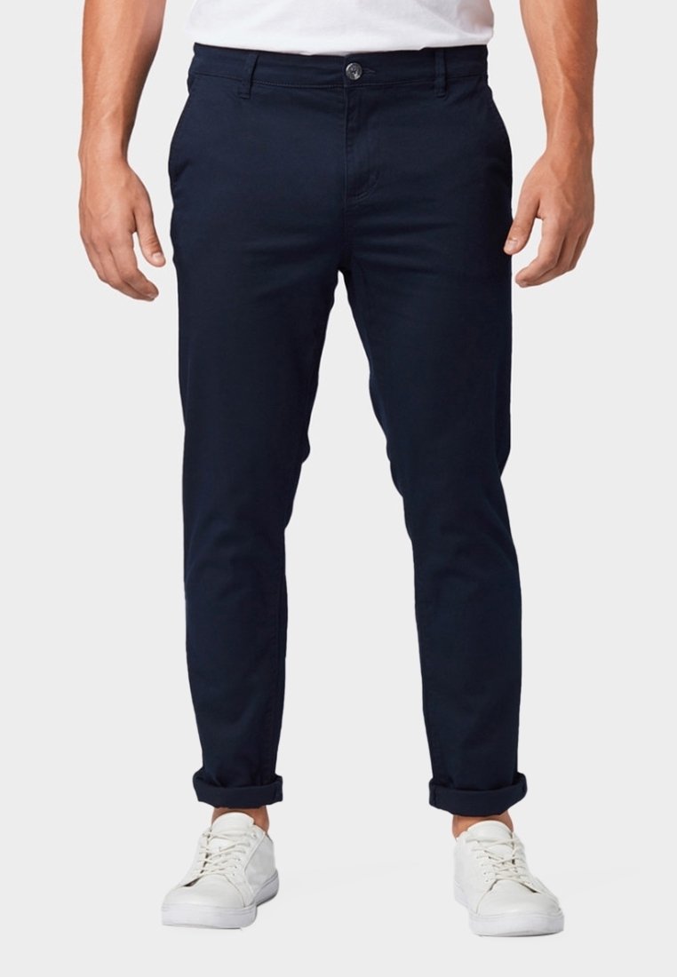 Homme portant un pantalon slim bleu marine retroussé et des baskets blanches, debout devant un fond uni.