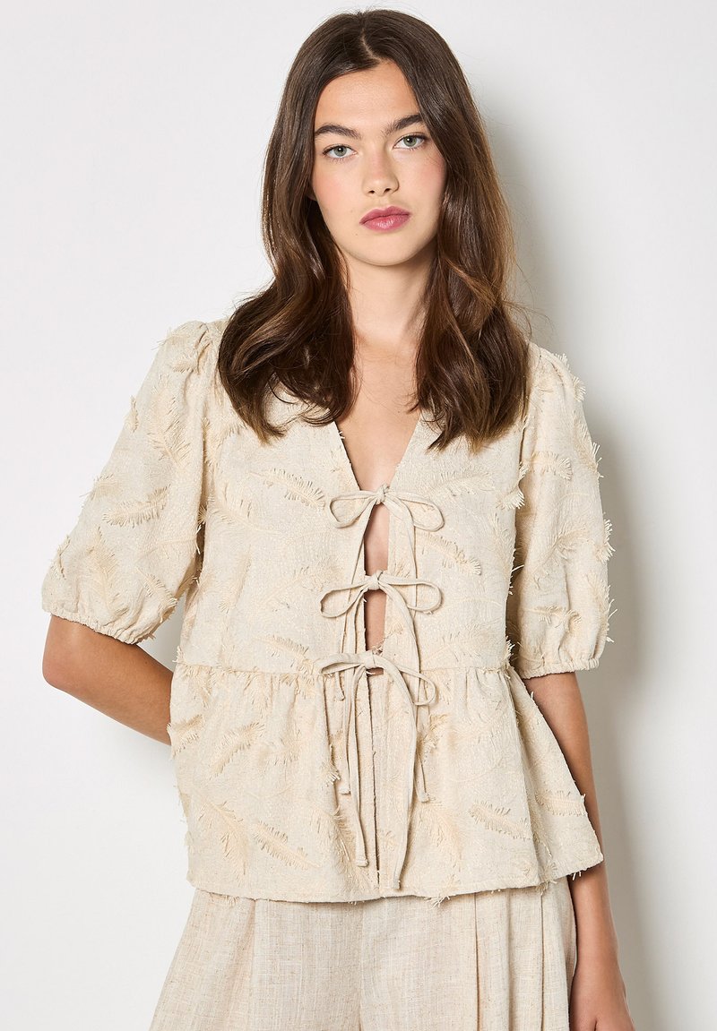 Jeune femme aux longs cheveux bruns portant un haut beige texturé avec trois liens à l'avant et des manches bouffantes jusqu'au coude.