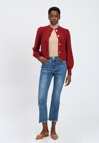 Rode gebreide cardigan met pofmouwen en gouden knopen, gecombineerd met hoog-waist blauwe jeans met een rafelige zoom en bruine ballerina's.