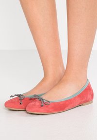 Korallfärgade mockaskor med lätt blå kant och grå rosett. Sulan är platt och skorna har en slät yta.