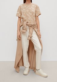 Beige T-shirt med hvide abstrakte bogstaver, parret med lyse, vide bukser og et draperet beige yderlag. Hvide sneakers.