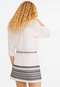 Mujer con cabello largo y rizado de color rubio que lleva una blusa blanca y una falda blanca con patrones de hojas negras y puntos, vista desde detrás.