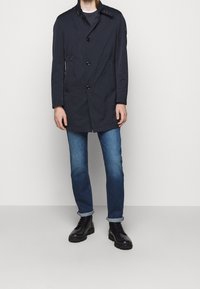 Námorně modrý kabát s knoflíkovým zapínáním a bočními kapsami, s výstužným límcem a knoflíkovým detailem na rameni, nošený přes modré džíny a černé boty.