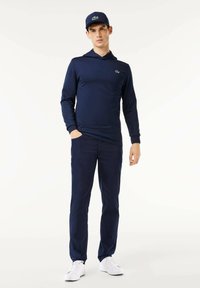 Marinblå sweatshirt med lång ärm och kenguruficka, tillsammans med matchande marinblå byxor. Vita sneakers och en marinblå keps med en grön logotypdetalj.