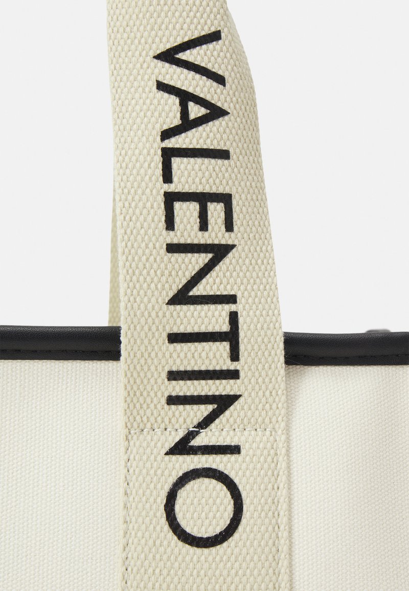 Bolso tote de lona con cuerpo crema y detalles en cuero negro. Presenta un destacado logo negro "VALENTINO" en la correa. Superficie texturizada.