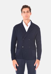 Cardigan doppiopetto blu navy, in tessuto a maglia testurizzato, con due tasche frontali, colletto a risvolto e bottoni. Indossato sopra una camicia bianca.