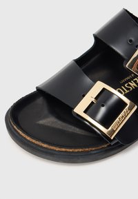 Μαύρα σανδάλια με γυαλιστερό φινίρισμα, που διαθέτουν δύο φαρδιές λωρίδες και χρυσή αγκράφα. Η σόλα είναι διαμ ορφωμένη και έχει καφέ σουέντ όψη.