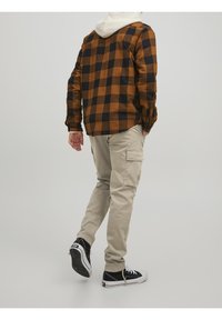 Brun och svart rutigt skjorta, beige cargo shorts, svarta sneakers. Texturerad tyg, avslappnad passform, synlig huva under skjortan.