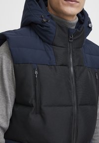 Padded vest featuring a navy hood, gray interior, and black lower section. Includes two zippered pockets and a front zipper closure.