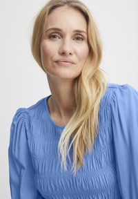 Lyseblå top med indsamlet tekstur, rund halsudskæring og puffede ærmer. Glat stof med subtil glans; håret er bølget og langt.