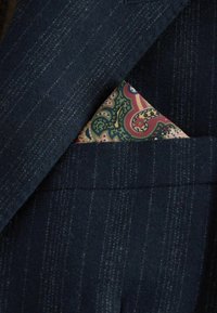 Sötétkék, testre szabott öltönyzakó texturált anyaggal és finom csíkozással. Egy mintás zsebkendő látható rózsaszín, zöld és barna színekben.