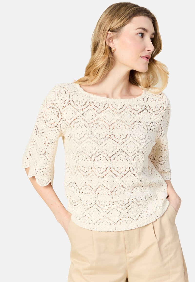 Top bianco in pizzo con motivo a diamante, scollo rotondo e maniche a tre quarti. Tessuto leggero con una finitura testurizzata.