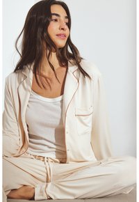 Crème-kleurige satijnen pyjama set met een knoopsluiting bovenkant, zakdetails en bijpassende drawstring broek met een gladde textuur en zachte glans.