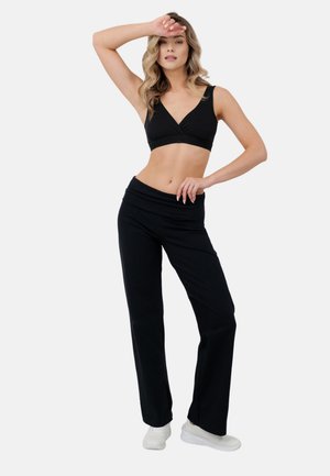 Kobieta stojąca, ubrana w czarny bezrękawnik crop top, czarne szerokie spodnie i białe sportowe buty na prostym jasnym tle.