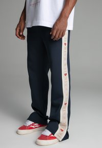 Navy track pants with contrasting cream side stripe, featuring orange branding. Soft material, fitted design, and zipper details at the hem.