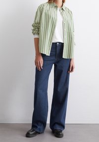 Camisa de botones a rayas verdes y blancas sobre una camiseta blanca, combinada con jeans de denim azul holgados y mocasines negros. Diseño minimalista.