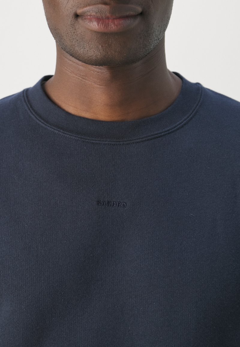 Marineblaue Sweatjacke aus weicher Baumwolle, mit geripptem Rundhalsausschnitt und geprägtem Logo "SANDRO" auf der Brust. Glatte Textur.