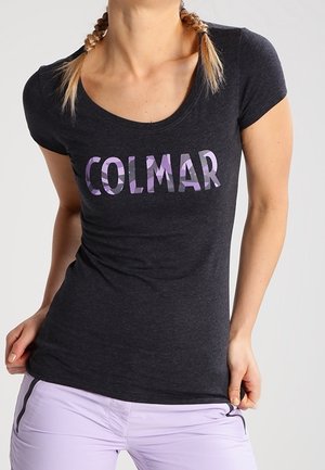 Femme portant un t-shirt noir cintré avec "COLMAR" en lettres violettes et un pantalon violet clair, tenant l'ourlet de sa chemise.