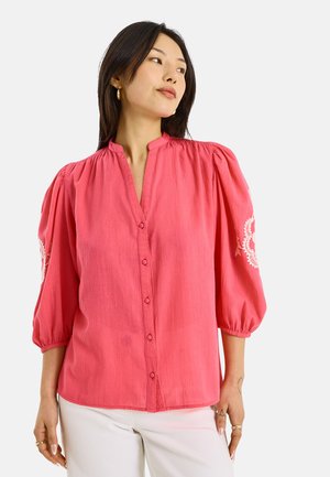 Femme portant un chemisier corail rose boutonné avec des manches trois-quarts bouffantes et une broderie blanche sur le haut des bras, associé à un pantalon blanc.