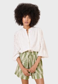 Blusa bianca ricamata con chiusura a bottoni e maniche a sbuffo, abbinata a pantaloni corte con coulisse a scacchi verdi. Tessuto morbido, motivi dettagliati.