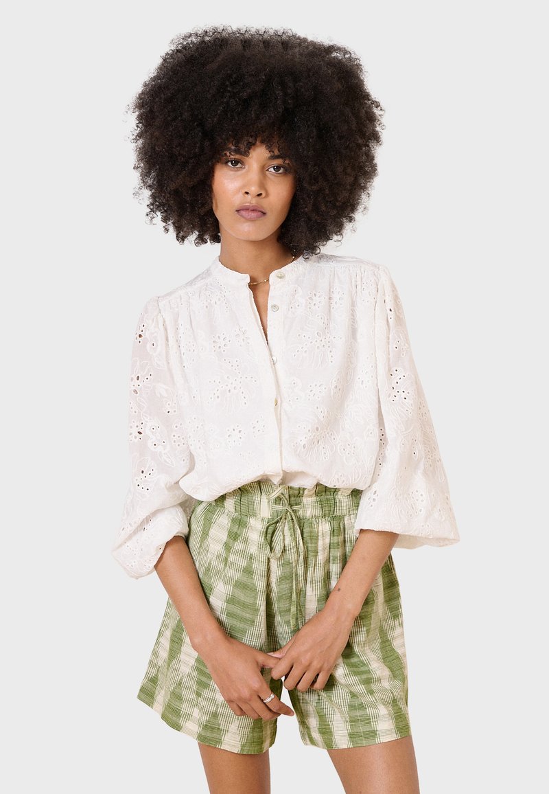 Blusa bianca ricamata con chiusura a bottoni e maniche a sbuffo, abbinata a pantaloni corte con coulisse a scacchi verdi. Tessuto morbido, motivi dettagliati.