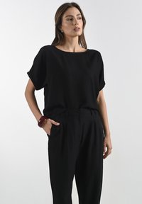Čierny krátky rukáv top s voľným strihom a širokým výstrihom, spárovaný so zúženými čiernymi nohavicami. Modelka nosí červený náramok.