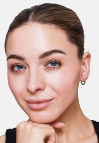 Primo piano di un volto con pelle liscia, capelli scuri e lisci, occhi azzurri e indossante un piccolo orecchino a cerchio in argento. Trucco semplice e naturale.