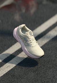 Lys beige og rosa joggesko med grå logo, snørebånd og polstret såle, svevende over en asfaltvei med hvite veimerkninger.