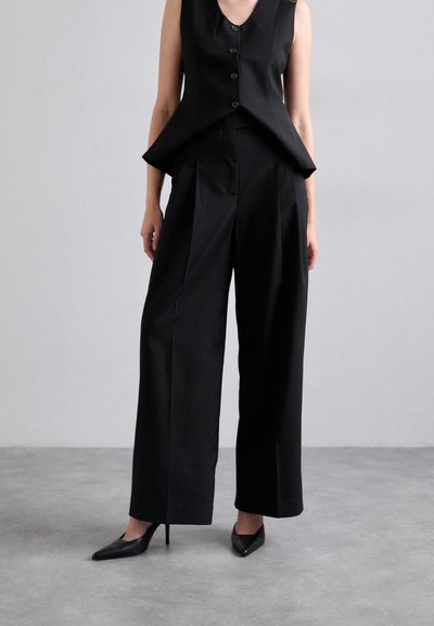 Zwarte mouwloze top met knoopsluiting, gecombineerd met hoge taille, wijde broek in een soepele, op maat gemaakte stof. Puntige zwarte hakken.