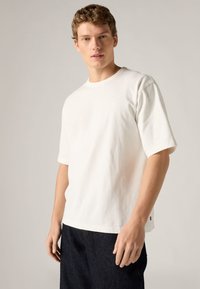 Λευκό βαμβακερό T-shirt με κοντά μανίκια, στρογγυλή λαιμόκοψη και μικρό λογότυπο στο μπροστινό μέρος. Συνδυασμένο με σκουρόχρωμο τζιν παντελόνι.