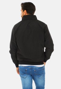 Giubbotto bomber nero con colletto alto, texture liscia, polsini aderenti e design leggermente corto. Indossato sopra jeans in denim blu.