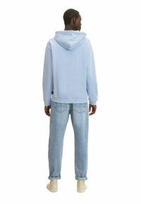 Felpa azzurra chiara con cappuccio regolabile, abbinata a jeans larghi in denim chiaro e sneakers beige, vista da dietro.