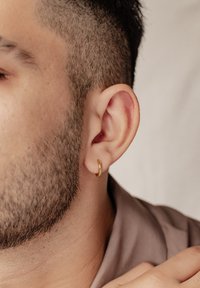 Orecchino a cerchio d'oro in primo piano su un orecchio di uomo, con capelli corti scuri e una sottile barba, su uno sfondo neutro morbido.