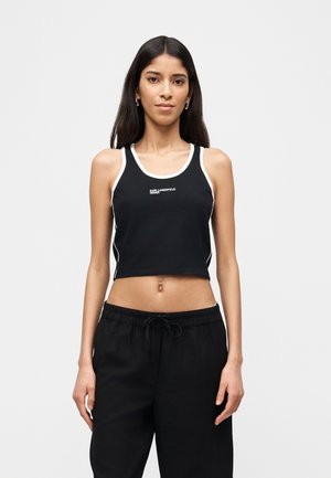 Jeune femme aux longs cheveux foncés portant un crop top noir à bordures blanches et un pantalon noir à cordon, faisant face à l'avant sur un fond blanc.