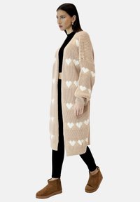 Długi, dziergany beżowy kardigan z kremowymi wzorami serc, z szerokimi rękawami i ściągaczami przy mankietach, noszony na czarnej bluzce i legginsach.
