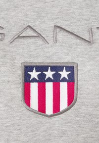 Sudadera gris con un logo de escudo bordado que presenta tres estrellas blancas sobre azul y franjas rojas y blancas debajo.