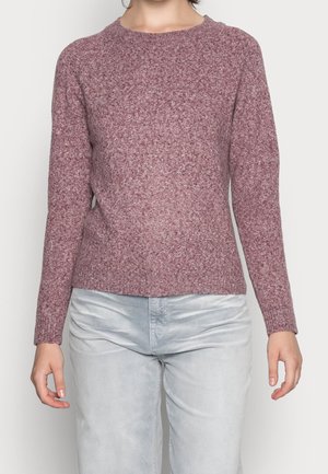Personne portant un pull mauve texturé à manches longues et un pantalon gris clair, debout devant un fond blanc uni.