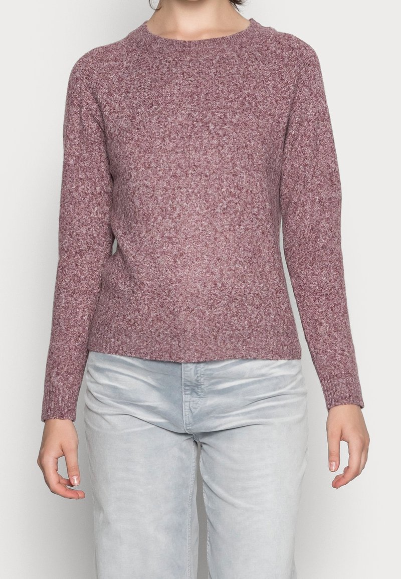 Personne portant un pull mauve texturé à manches longues et un pantalon gris clair, debout devant un fond blanc uni.