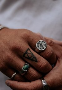 Silverringar med intrikata mönster och en grön sten, bärs på en hand med tatueringar och texturerad hud.