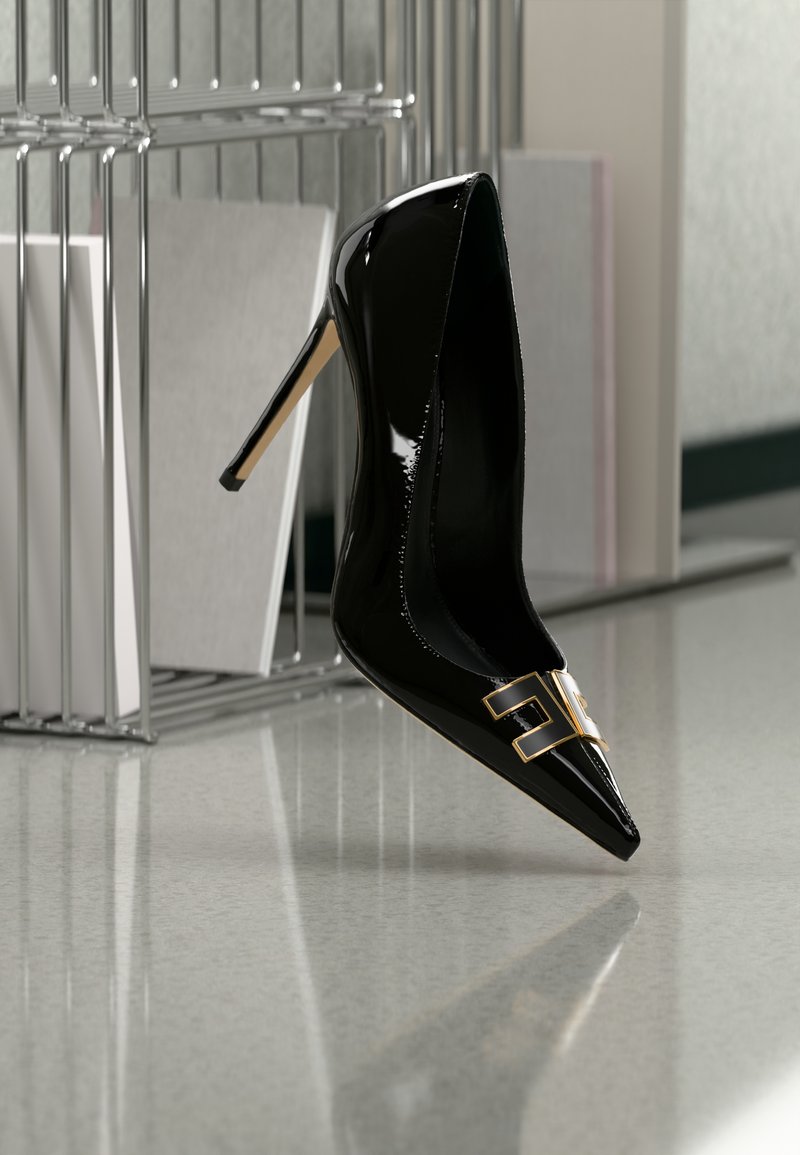 Μαύρο στιλέτο από δέρμα με φινίρισμα πατέντα και μυτερή μύτη, με διακοσμητικό χρυσό στοιχείο στο μπροστινό μέρος, τοποθετημένο σε ανακλαστική επιφάνεια.